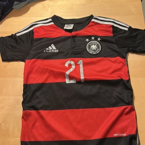 Tysklands Reus #21 Adidas fotbollströja - Snygg Tyskland fotbollströja från Adidas med Reus och nummer 21 på ryggen. Tröjan har breda svarta och röda ränder, silvriga detaljer och tre stjärnor ovanför klubbmärket. Kortärmad modell i lätt och ventilerande climacool-material.