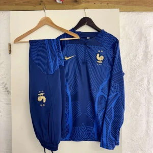 Frankrike långärmad tracksuit Nike storlek M - Blå långärmad fotbollströja från Nike med Frankrikes landslagsemblem och två stjärnor broderade i guld. Tröjan har ett grafiskt mönster i olika blå nyanser och dragkedja vid halsen. Perfekt för dig som älskar fotboll och vill sticka ut på planen.