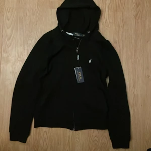 Svart hoodie Polo Ralph Lauren - Svart hoodie från Polo Ralph Lauren med broderad logga på bröstet. Slim fit-modell med huva och dragkedja framtill. Klassisk och stilren design, perfekt för dig som gillar en clean look. Materialet känns mjukt och bekvämt.