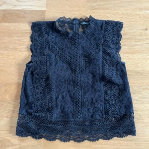 Svart spets blus med hög krage - Supersnygg svart blus  i spets med hög krage och ärmlös design. Blusen har vackra blommiga spetsdetaljer, vågiga kanter och en liten öppning med knapp i nacken. Perfekt för dig som gillar romantisk och trendig stil.