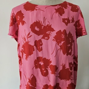 Rosa blommig blus från Jascha Stockholm  - Superfin blus från Jascha i rosa med stora röda blommor. Modellen är rak med korta ärmar och rund halsringning. Tillverkad i mjuk viskos som känns skön mot huden. Perfekt för dig som gillar färg och mönster!