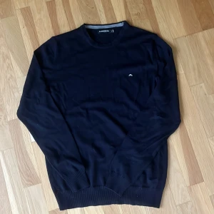 Mörkblå stickad tröja J.Lindeberg - Snygg mörkblå stickad tröja från J.Lindeberg med rund halsringning och diskret logga på bröstet. Tröjan har ribbade muddar vid ärmslut och nederkant, perfekt för en stilren look. Passar dig som gillar klassisk och enkel stil.