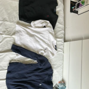 3 långärmade tröjor Polo Ralph Lauren - Tre långärmade tröjor från Polo Ralph Lauren. En svart sweatshirt med röd logga, en vit och en marinblå ribbad tröja med broderad logga. Alla har klassisk passform och rund hals. Perfekta för lager-på-lager och stilren vardagslook.