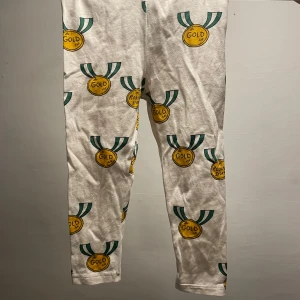 Mini Rodini medaljleggings, vit/gul - Snygga vita leggings från Mini Rodini med coolt medaljmönster i gult och grönt. Gjorda i mjuk bomull med lite stretch för skön passform. Perfekta för dig som gillar lekfull stil och vill sticka ut lite extra. Fläckar syns på bilder 