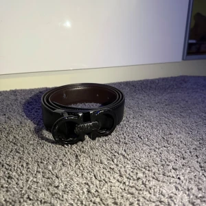 Svart och brunt skärp från Salvatore Ferragamo - Snyggt bälte från Salvatore Ferragamo i svart och brunt skinn med ikoniskt dubbel-Gancini spänne i svart metall. Bältet har stilren design och präglad logga på insidan. Perfekt för att lyfta din outfit med en lyxig touch. (Lite söndrig på insidan, inget som märks) 