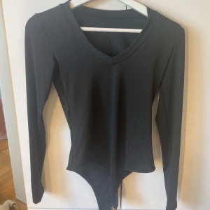 Svart långärmad bodysuit med v-ringning - En stilren svart bodysuit med lång ärm och snygg v-ringning. Tight passform som sitter på plats hela dagen. Perfekt att styla med jeans eller kjol för en clean look. Gjord i mjukt och stretchigt material för maximal komfort.