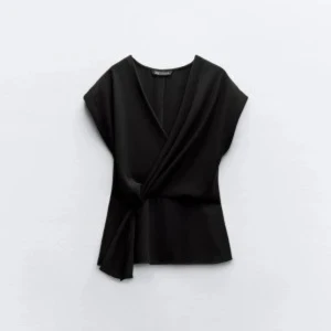 Svart omlottopp med knyt - Säljer en svart omlottopp med snygg knytning i sidan och v-ringad hals. Toppen har korta ärmar och en stilren, lite lös passform. Perfekt att matcha med jeans eller kjol för en cool look.