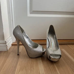 Glittriga silverfärgade pumps med platå - Snygga pumps i glittrigt silver med hög klack och platåsula. Skorna har öppen tå och rundad form framtill. 