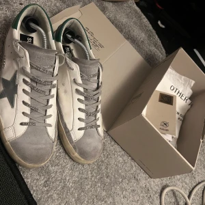 Golden Goose vita sneakers med grön detalj - Golden Goose sneakers i vitt skinn med grå mockadetaljer och grön häl. Klassisk stjärna på sidan och grå snörning med text. Snygg vintage-look och platt sula. Kommer med originalkartong och dustbag. helt äkta köpte från minto för ca 6000