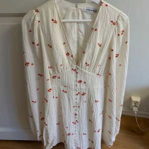 Vit blommig klänning, Adoore - Supersöt vit kortklänning från Adoore med små röda blommor, v-ringning och knappar framtill. Långärmad med lätt struktur i tyget och markerad midja. Så fin till midsommar💗