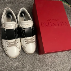 Valentino Garavani Rockstud sneakers - Snygga Valentino Garavani Rockstud sneakers i vitt skinn med svart band över vristen och ikoniska nitar baktill. Klassisk rund tå, platt sula och snörning. Kommer med originalkartong, extra skosnören och dustbags. Perfekta för dig som vill sticka ut med lyxig streetstyle. helt äkta, hör av dig om frågor