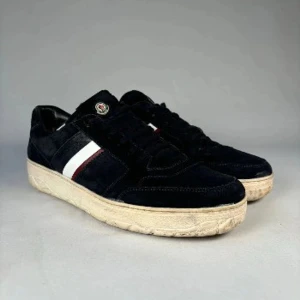 Moncler svarta sneakers i mocka - Snygga svarta sneakers från Moncler i mocka med vita och röda detaljer på sidan. Skorna har en klassisk låg profil, rund tå och snörning. Tjock vit sula med Moncler-logga på plösen ger en exklusiv vibe. Perfekta för dig som gillar stilrena och sportiga skor.