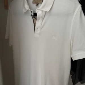 Vit Burberry pikétröja  - Säljer en klassisk vit pikétröja från Burberry i 100% bomull. Tröjan har korta ärmar, krage och knäppning framtill med den ikoniska Burberry-detaljen vid knappslån. Perfekt för en clean och stilren look. I mycket bra skick! Hör av er vid frågor och funderingar, mvh.