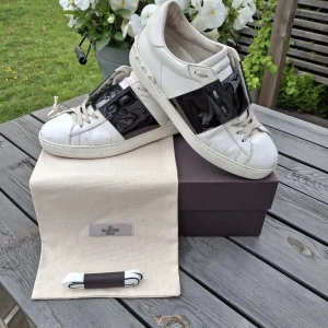 Valentino Garavani Open sneakers vit/svart - Snygga Valentino Garavani Open sneakers i vitt skinn med en bred svart lackdetalj över mitten. Klassisk rund tå, vita snören och coola nitar baktill på sulan. Befintligt skick, se bilder. Hör av dig vid frågor eller liknande. Priset är inte hugget i sten.