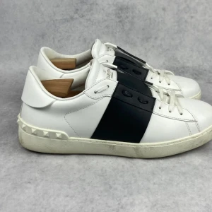 Valentino Garavani Open sneakers vit/svart - Snygga Valentino Garavani Open sneakers i vitt skinn med bred svart läderdetalj över snörningen. Klassisk rund tå, platt sula och ikoniska nitar baktill på hälen. Perfekt för dig som gillar stilrena och lyxiga sneakers med en edgy twist.