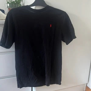 Svart t-shirt från Polo Ralph Lauren - Svart t-shirt från Polo Ralph Lauren i bomull med klassisk röd broderad logga på bröstet. Enkel och stilren design med rund hals och korta ärmar. Perfekt för dig som gillar en clean och tidlös look.