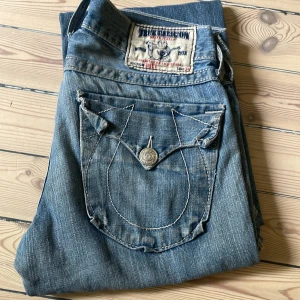 True Religion jeans blå denim - Snygga blå jeans från True Religion med ikoniska kontrastsömmar och stora fickor med knappdetaljer. Klassisk femficksmodell i tvättad denim med slitningar framtill för en cool look. Perfekta för dig som gillar streetstyle och vill sticka ut.