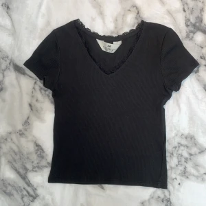 Svart ribbad topp med spets från H&M - Svart ribbad topp från H&M med korta ärmar och v-ringning. Fina spetsdetaljer runt halsringningen ger en extra touch. Toppen är figurnära och har en enkel, stilren look som passar till många outfits.