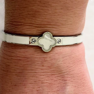 Silverfärgat armband med vit detalj - Stilrent armband i silverfärg med vit emalj och gnistrande stenar på sidorna. Framtill sitter en fyrklöverformad detalj med pärlemorskimrande yta och  strass runt kanten. Perfekt accessoar för att lyfta din look.