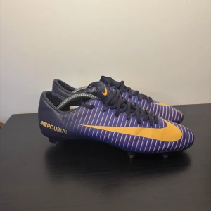 Nike Mercurial blå-orange fotbollsskor storlek 46 - Säljer ett par Nike Mercurial fotbollsskor i mörkblått med orange swoosh och tunna randiga detaljer i rosa och blått. Skorna har snörning och metall-dobbar för bästa grepp på planen. Snygg, sportig design som sticker ut på matchen.