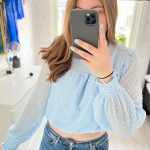 Ljusblå blus med puffärmar  - Supersöt ljusblå blusen prislapp kvar. Med smock upptill, transparenta puffärmar och diskret prickigt mönster. Croppad modell med volangdetaljer vid axlar och ärmslut. Perfekt för dig som gillar romantisk och trendig stil. Svalt och skönt material som tyvärr aldrig kommit till användning. Storlek S
