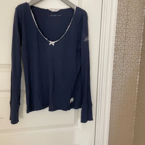Mörkblå ribbad topp Odd Molly - Supersöt mörkblå långärmad ribbad topp från Odd Molly med v-ringning, spetskant och liten rosett framtill. Skön stretchig bomullskvalitet som sitter snyggt och har extra långa ärmar. Perfekt för dig som gillar en feminin och chill stil. Lite urtvättad men annars i fint skick. Passar dig som är storlek small till medium. 