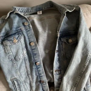 Ljusblå jeansjacka - Säljer en klassisk ljusblå jeansjacka från &Denim i storlek 34. Jackan har silverfärgade knappar, två bröstfickor med lock och knapp samt snygga sömdetaljer!