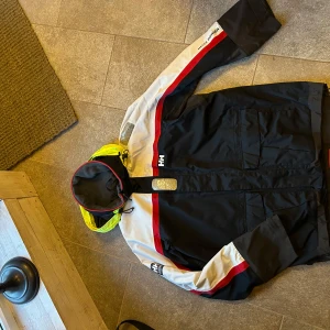 Skaljacka Helly Hansen - Sparsamt använd skaljacka från Helly Hansen. Vind och vattentät. Strl XL. Lättare slitage vid armarnas nedre del, se bild 4. Marinblå med röda, vita och gula detaljer.