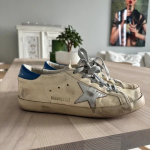 Golden Goose Superstar sneakers med silverdetaljer - Golden Goose Superstar sneakers i beige skinn med silverfärgad stjärna på sidan, glittriga silverfärgade snören och blå hälkappa. Perfekta för dig som gillar en cool och avslappnad stil. Golden goose i limited edition . Skorna har färgerna blå beige och en silvrig stjärna med silvriga skosnören till. Skorna är väl använda men inte något som märks då det ska ge en sliten känsla. Skorna köptes i Frankrike som är för 660 euro har tyvärr inget kvitto då det köptes i butik. Dustbag medföljer 