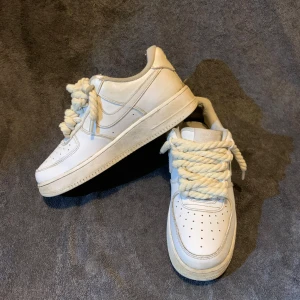 Nike Air Force 1 vita sneakers - Klassiska Nike Air Force 1 sneakers i helvitt skinn med chunky, rep-liknande snören. Låg modell med perforerad tå och rundad sula. Perfekta för dig som gillar en clean och tidlös look med en twist på snörningen.