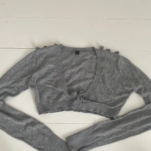 Grå bolero med rosetter från Bloch - Superfin grå croppad kofta från Bloch med långa ärmar med hål för tummen och söta rosett detaljer längs ärmarna och framtill. Den är gjord av 100% bomull och köpt i dansbutiken i Stockholm. Väldigt bra skick och fantastisk kvalitet!