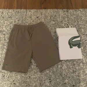 Beige shorts från Lacoste med snörning - Snygga beige shorts från Lacoste med klassisk krokodillogo på benet. De har elastisk midja med snörning och är tillverkade i mjuk bomull, perfekta för chill dagar. Passar dig som gillar enkel och stilren look. Storlek M men, passar som Storlek S 