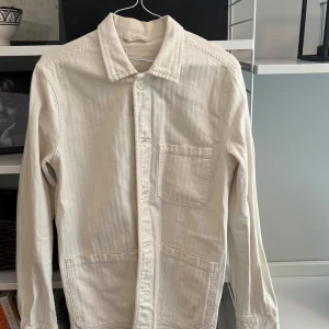 Vit overshirt i bomull - Stilren vit overshirt i bomull med klassisk krage, tre fickor framtill och knäppning. Perfekt att slänga över en t-shirt för en avslappnad look. Långärmad och enkel design som funkar till det mesta.