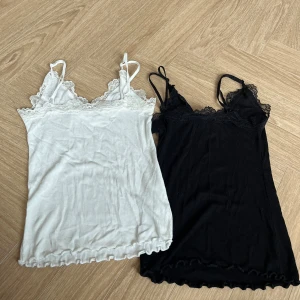 2 ribbade spetslinnentoppar Amisu XXS - Två ribbade linnetoppar från Amisu i storlek XXS, en vit och en svart. Båda har tunna axelband, spetskant upptill och vågig kant nertill. Perfekta att styla lager på lager eller med jeans för en chill look.