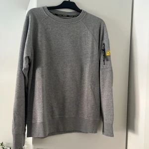 Grå sweatshirt från Acqua Limone - Snygg grå sweatshirt från Acqua Limone med klassisk citronsymbol i gult på ärmen och nacken. Tröjan har lång ärm, rund hals och en cool ficka med dragkedja på ärmen. Perfekt för dig som gillar stilrena och bekväma plagg.