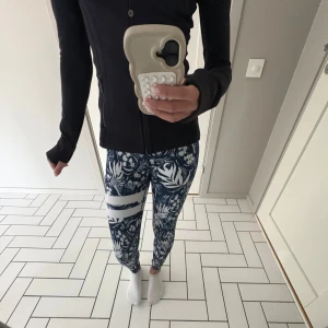 Blommiga leggings från Stronger XS - Snygga leggings från Stronger i storlek XS med mörkblå och vita blommönster samt två vita ränder på ena benet. Hög midja och tight passform. Tillverkade i återvunnen polyester och elastan, perfekta för träning eller chill.