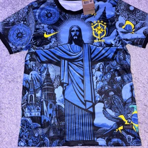 Nike Brasil Jesus fotbollströja  - Unik blå Brasil-fotbollströja från Nike med grafiskt tryck av Kristusstatyn, kyrkor och brasilianska symboler. Gula detaljer som Nike-logga och Brasil-märke. Tillverkad i lätt, ventilerande polyester med svart krage och korta ärmar. Perfekt för dig som vill sticka ut på planen.