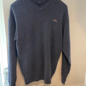 Mörkgrå stickad tröja från GANT - Säljer en mörkgrå stickad tröja från GANT med v-ringad hals och broderad GANT USA-flagga på bröstet. Tröjan har lång ärm och klassisk passform, perfekt för lager-på-lager under kyliga dagar.