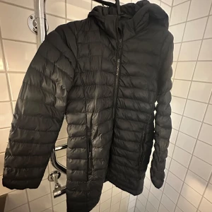 Svart dunjacka med huva EVEREST - Säljer en svart dunjacka med huva och dragkedja framtill. Jackan har quiltad design, två fickor med dragkedja och lång ärm. Perfekt för kyliga dagar och enkel att matcha med allt. Snygg och funktionell för dig som vill hålla värmen. Från Everest 