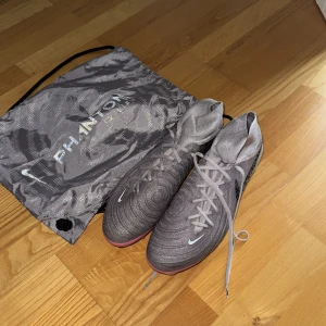 Nike Phantom GX Luna Elite grå fotbollsskor s47. - Nike Phantom GX Elite fotbollsskor i grått med svarta detaljer och skimrande sula i rosa och blått. Skorna har hög strumpliknande krage, snörning och mönstrad ovandel. Perfekta för dig som vill sticka ut på planen. Medföljer originalpåse. Storlek 46!