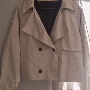 Beige croppad trenchcoat med knappar - Snygg beige croppad trenchcoat med stora slag och dubbelknäppning framtill. Jackan har långa ärmar med detalj vid ärmslut och två fickor framtill. Den är inte så jätte varm så den är perfekt att ha på sig den under sommaren när de lite kallt!🌸 Säljer den då jag fick den e fel storlek😮‍💨