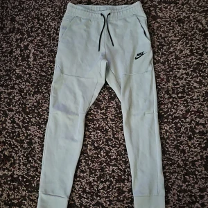 Ljusgrå mjukisbyxor från Nike - Snygga ljusgrå mjukisbyxor från Nike med svart logga på vänster ben. Byxorna har dragsko i midjan, ribbade muddar vid bensluten och praktiska fickor. Tillverkade i mjukt bomullsmaterial för maximal komfort. Perfekta för chill eller träning.