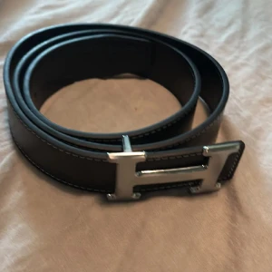 Svart H skärp med H-spänne - Snyggt svart skärp från H med ikoniskt H-format spänne i silverfärg. Bältet är gjort i slätt skinn med vita kontrastsömmar längs kanterna. Perfekt accessoar för att lyfta din outfit med en lyxig touch.