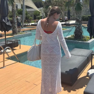 Vit virkad långklänning med öppen rygg - Superfin vit virkad långklänning med öppen rygg och lång ärm. Perfekt för sommardagar vid poolen eller stranden. Klänningen har ett snyggt mönster och är lätt transparent, vilket gör den extra somrig och chill.
