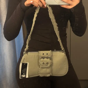 Beige axelväska med fransar och bälte - Trendig beige axelväska i bomull med fransade kanter och coolt bältedetalj framtill. Väskan har dragkedja upptill och en bred axelrem med råa kanter för en edgy look. Perfekt för att lyfta din outfit med en unik touch.