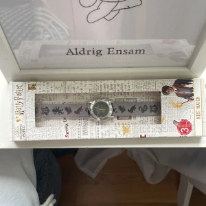 Harry Potter barnklocka med tygarmband - Grym barnklocka med Harry Potter-tema! Klockan har ett silverfärgat metallboett och ett grått tygarmband med svarta siluetter av djur från Hogwarts. Urtavlan har Hogwarts-vapnet och Harry Potter-loggan. Perfekt accessoar för alla unga Potterheads!