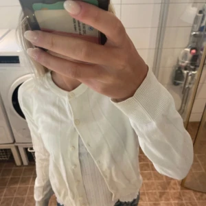 Vit kofta från H&M  - Säljer en klassisk vit kofta från H&M i mjuk bomull. Den har rund halsringning, långa ärmar och knäppning framtill med små vita knappar. Perfekt att slänga över en topp eller klänning för en clean och enkel look.