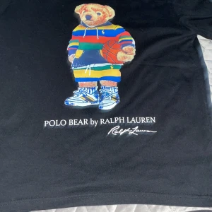 Svart Polo Bear t-shirt från Ralph Lauren - Svart t-shirt från Ralph Lauren med ikoniska Polo Bear-trycket framtill. Björnen har färgglada kläder och håller en basketboll. T-shirten har rund hals och klassisk passform. Perfekt statement-plagg för dig som gillar streetstyle.
