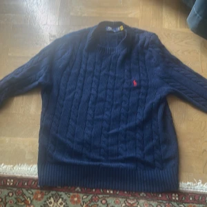 Mörkblå kabelstickad tröja Polo Ralph Lauren - Kabelstickad mörkblå tröja från Polo Ralph Lauren med klassisk rund hals och röd broderad logga på bröstet. Tröjan har långa ärmar och ribbade muddar vid ärmslut och nederkant. Perfekt för dig som gillar preppy och tidlös stil.