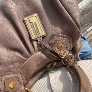 Brun skinnväska Marc by Marc Jacobs - Säljer en snygg brun handväska från Marc by Marc Jacobs i äkta italienskt skinn. Väskan har guldiga metalldetaljer, coolt märke framtill och justerbart handtag. Perfekt storlek för vardagens essentials och ger en lyxig touch till din outfit.
